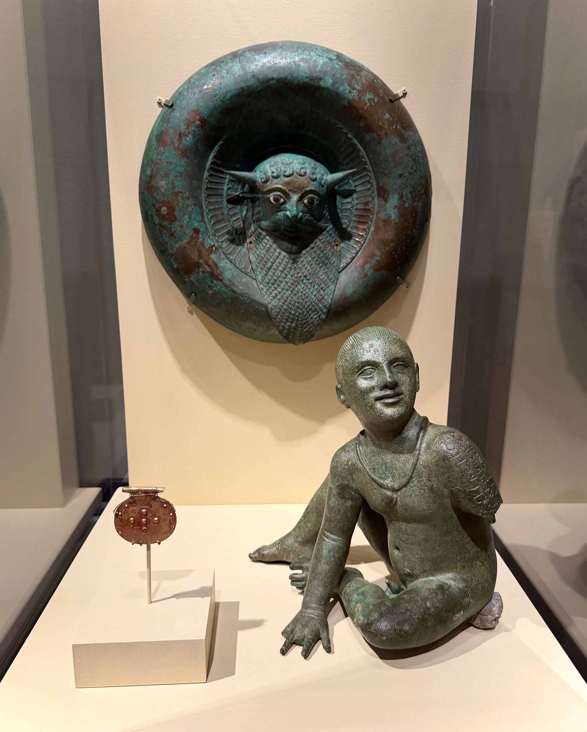 Three Etruscan Bronze Objects: Bulla, Round Boss with Head of Acheloös, and Votive Statue of a Seated Boy (“Putto Carrara”); Late 8th–3rd century BC. The Etruscans: From the Heart of Ancient Italy. Legion of Honor, SF.